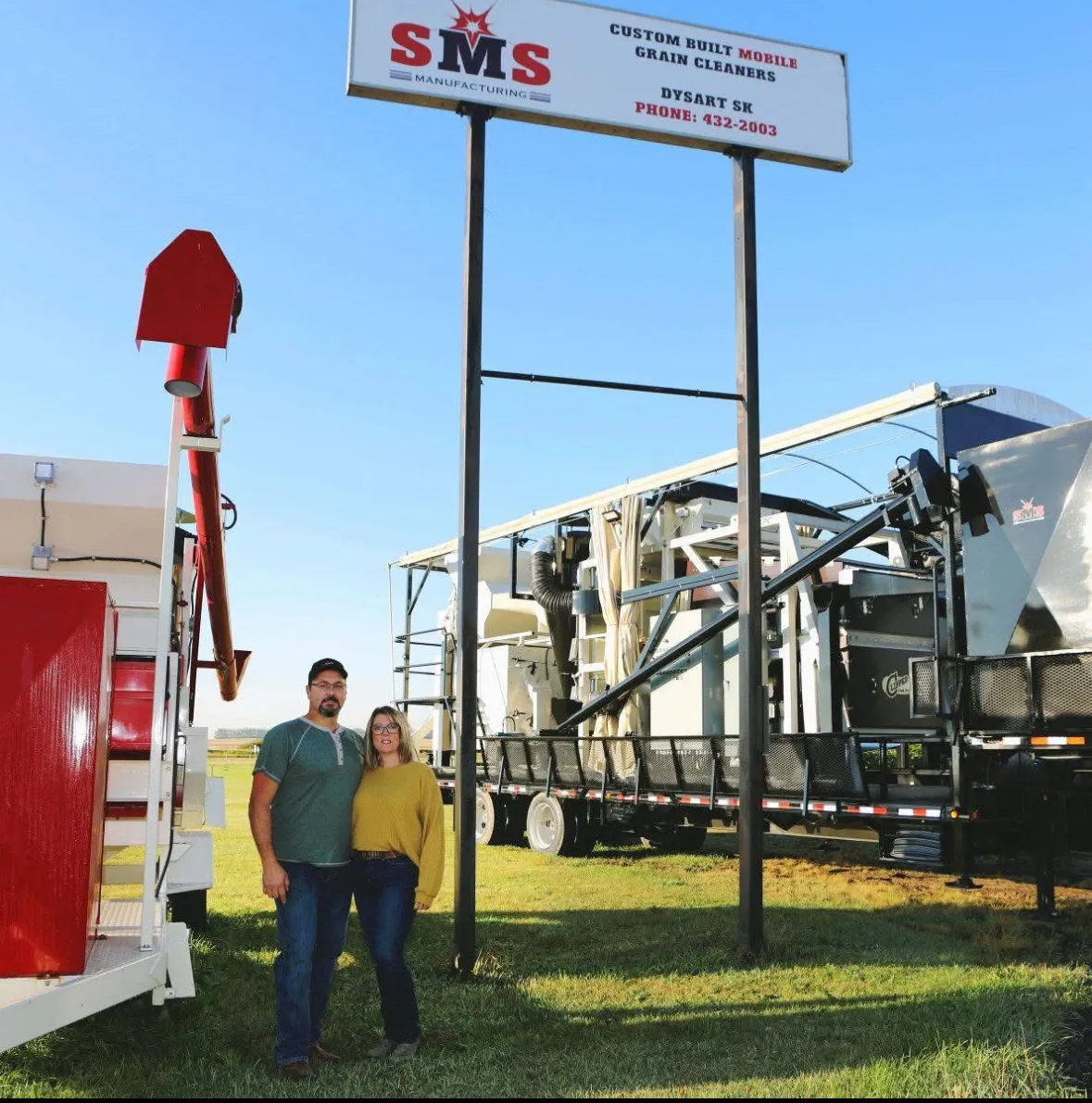 Two people standing beneath the SMS Manufacturing roadside sign in Dysart, with cleaners on either side
